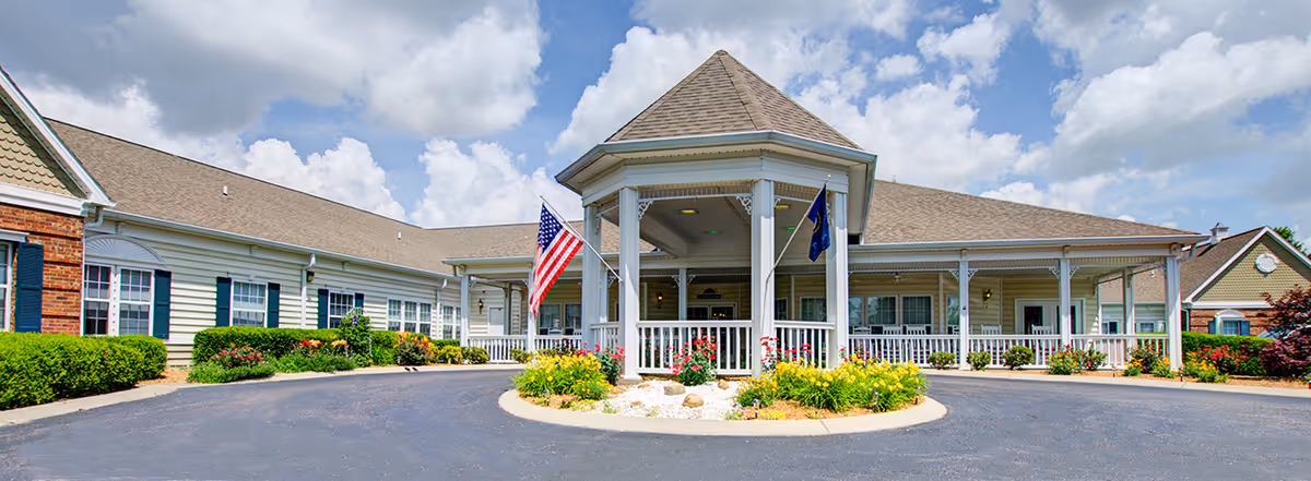 Front exterior view of The Bungalows at Mayfield facility featuring a covered entrance with white columns, American and state flags, surrounding landscaped flower beds, and a circular driveway under a partly cloudy sky.