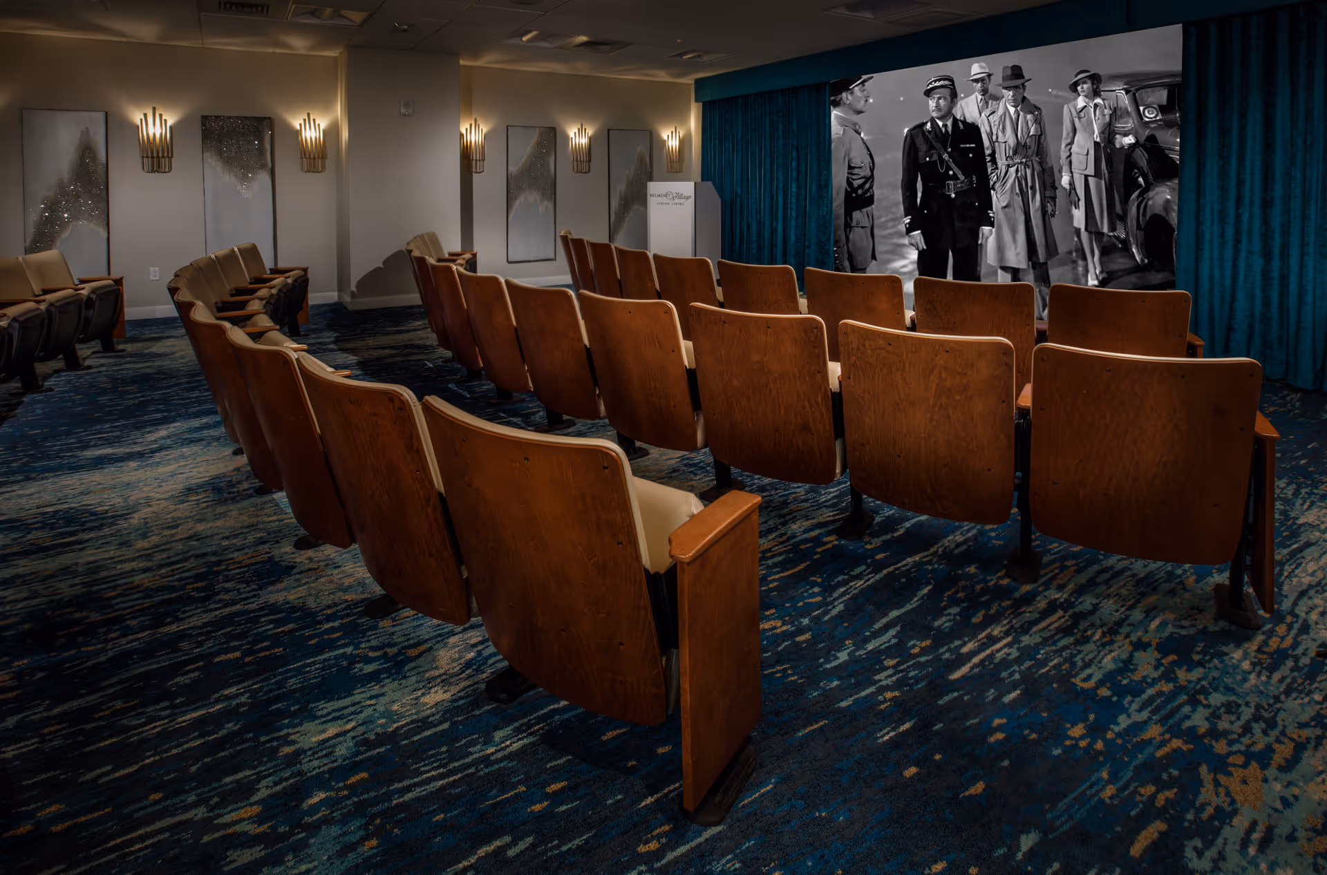 A small theater room with rows of wooden seats facing a large screen showing a black and white movie scene. The room has blue patterned carpet, wall sconces, and decorative wall panels.
