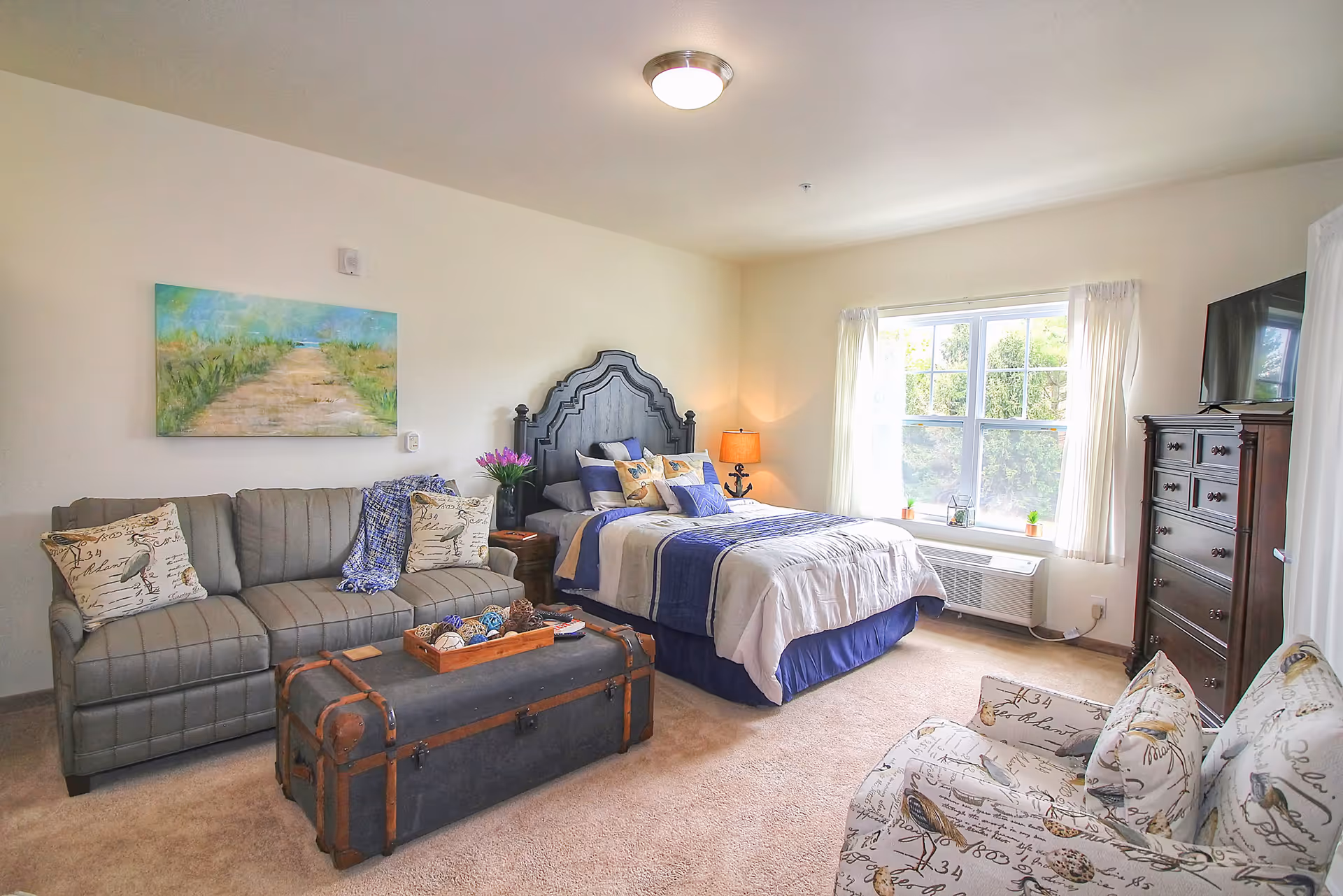 Well-lit furnished bedroom with a bed, sofa, trunk-style coffee table, armchair, large window and dresser.