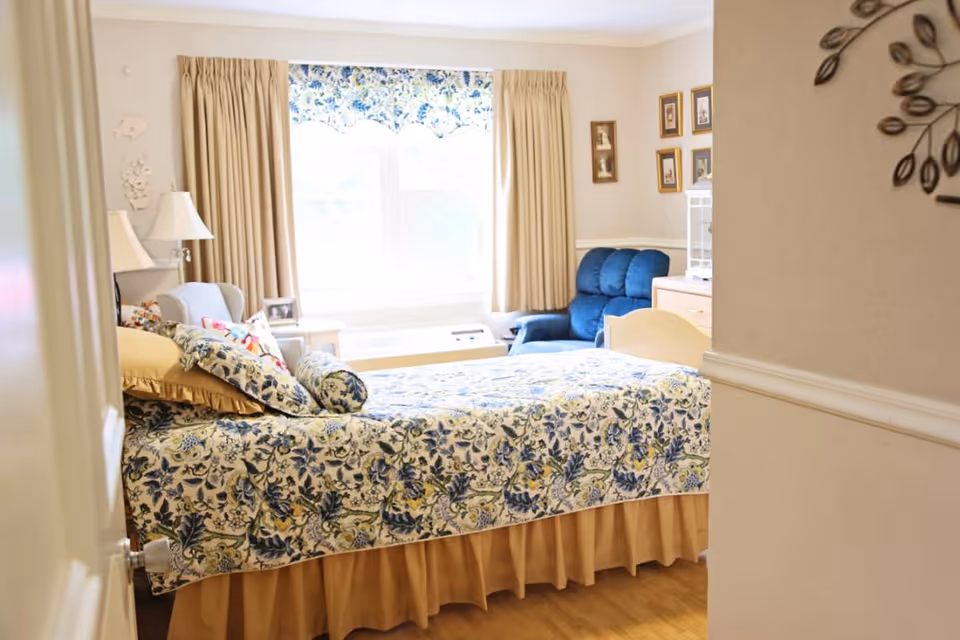 A cozy bedroom with a bed covered in a floral patterned bedspread in shades of blue and beige. The room has beige curtains and a matching valance over a large window. There is a blue armchair and a light blue chair near the window, along with a small side table and a lamp. The walls are decorated with framed pictures and a metal wall art piece.