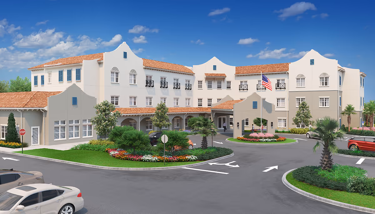 Front exterior of a Mediterranean-style three-story senior living building with a drive-up roundabout, landscaped islands, and parked cars under a blue sky.