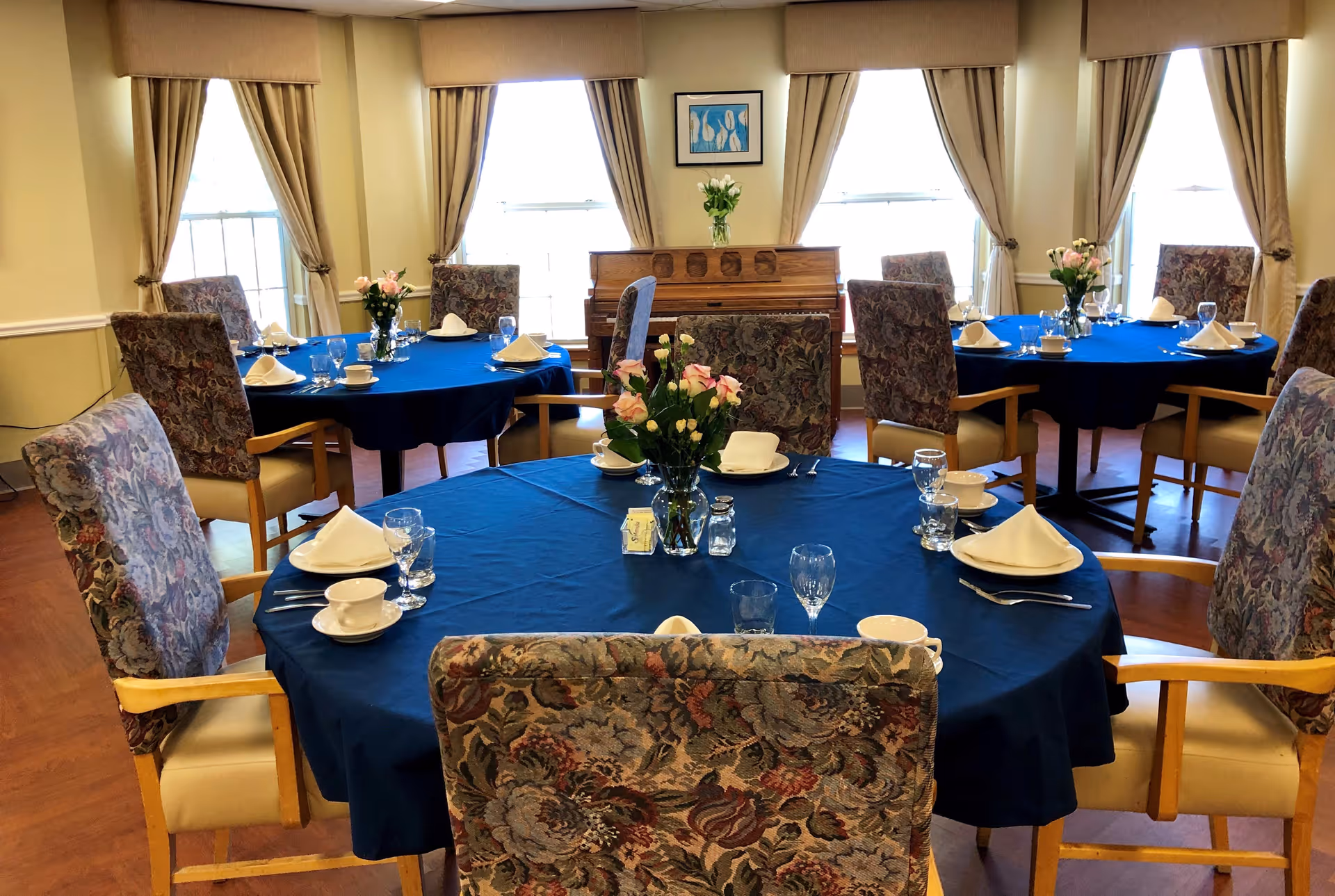 A dining room with round tables covered in blue tablecloths, each set with white napkins, cups, glasses, and silverware. The chairs have floral patterned upholstery and wooden armrests. There are three large windows with beige curtains, a piano against the wall, and a framed picture above the piano. Flower vases are placed on each table.