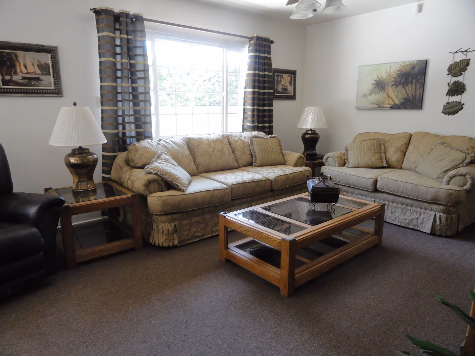 A living room with two beige patterned sofas, a wooden coffee table with glass top, two side tables each with a lamp, a large window with striped curtains, and framed artwork on the walls.