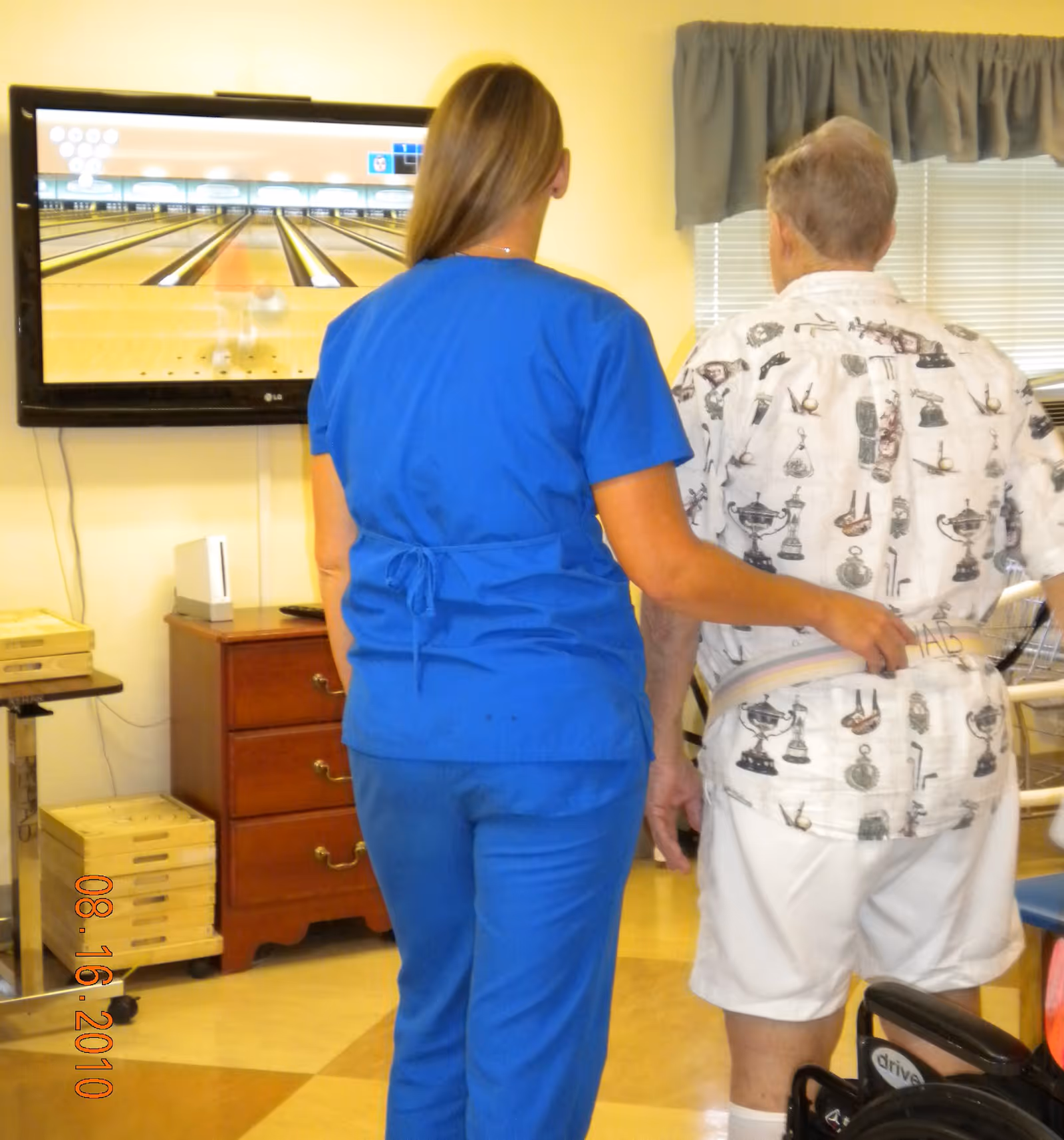 A caregiver in blue scrubs assists an elderly man wearing a patterned shirt and white shorts as they stand in front of a television screen displaying a virtual bowling game in a room with yellow walls and a window with blinds and a valance.