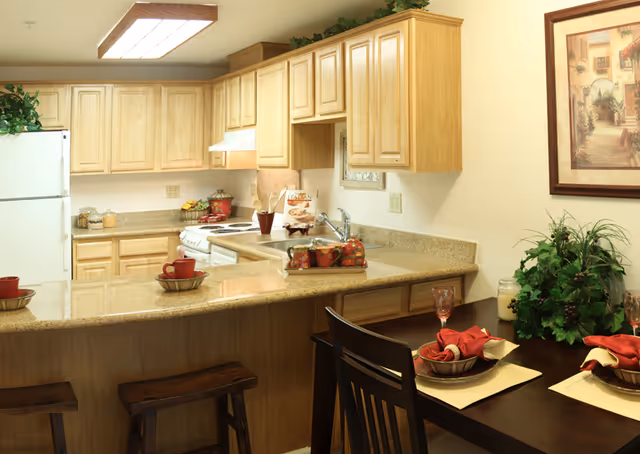 A well-lit kitchen with light wood cabinets, a white refrigerator, stove, and dishwasher. The kitchen has a granite countertop with two wooden stools at a breakfast bar. Adjacent to the kitchen is a dining table set with red napkins, plates, and glasses, along with a green plant centerpiece and a framed painting on the wall.