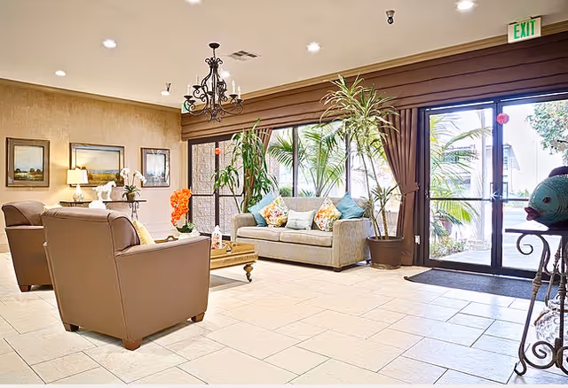 A bright and welcoming living room area with beige tiled floors, a beige sofa with colorful cushions, two brown armchairs, a wooden coffee table with decorative items, several potted plants, framed artwork on the walls, and large glass doors letting in natural light.