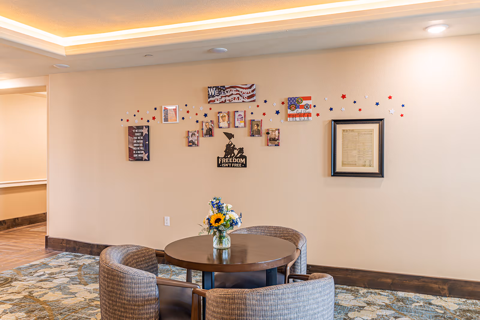 Small seating area with a round table, four upholstered chairs, a vase of flowers, and patriotic wall decorations in a senior living facility.