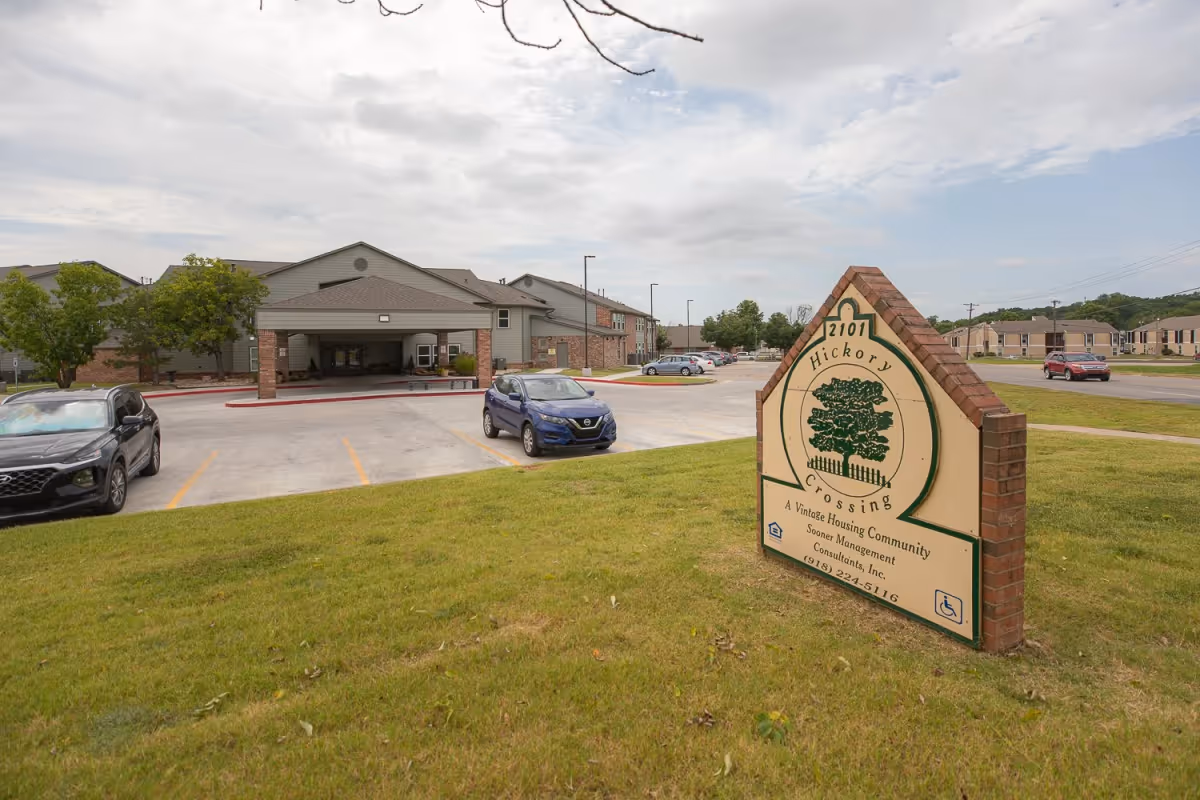 Exterior view of Hickory Crossing senior living facility with a brick sign on the lawn and cars in the parking lot.