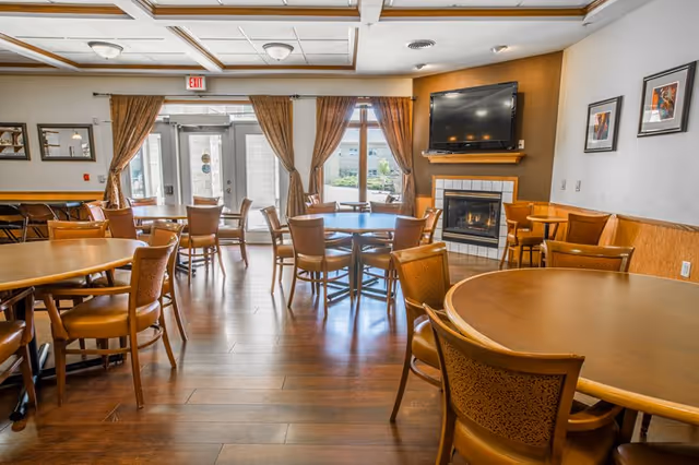 A well-lit dining room with multiple round wooden tables and chairs arranged neatly. The room features large windows with brown curtains allowing natural light to enter. A fireplace with a mounted flat-screen TV above it is visible on one wall, along with framed artwork. The floor is wooden, and the ceiling has decorative beams.