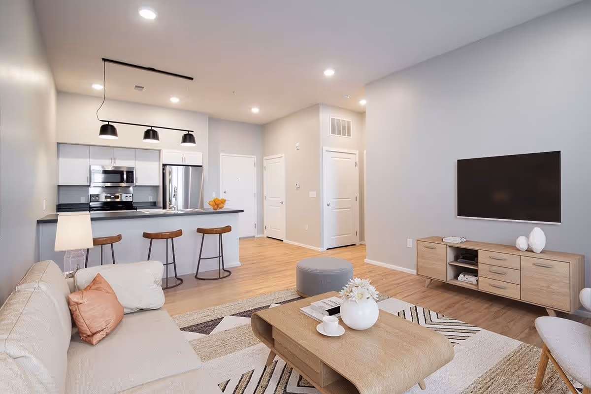A modern open-concept living room and kitchen area with light wood flooring and neutral-colored walls. The living room features a beige sofa with cushions, a wooden coffee table with decorative items, a gray ottoman, and a wall-mounted flat-screen TV above a wooden media console. The kitchen has white cabinetry, stainless steel appliances including a refrigerator and oven, a countertop with three wooden bar stools, and three black pendant lights hanging above the counter.
