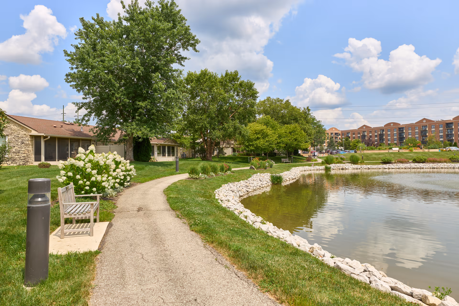A paved walking path curves alongside a small pond bordered with white rocks. There are green grassy areas, trees, and bushes with white flowers along the path. A wooden bench is placed near the path next to a lamp post. In the background, there are low buildings and a larger multi-story building under a partly cloudy blue sky.