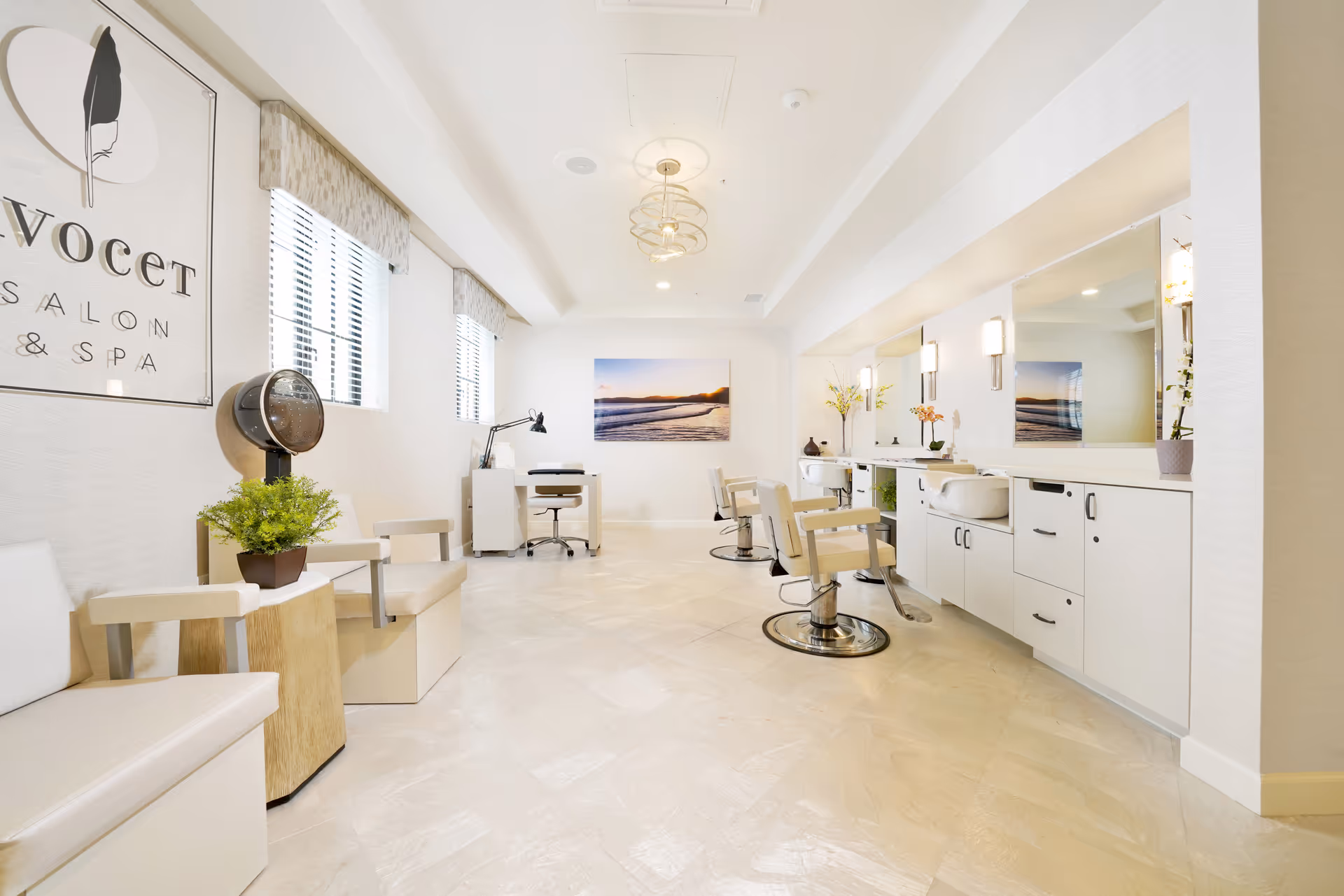 Bright and modern salon room with white walls and flooring, featuring salon chairs in front of large mirrors, a hair dryer chair, a manicure table with a lamp, and decorative plants and artwork on the walls.