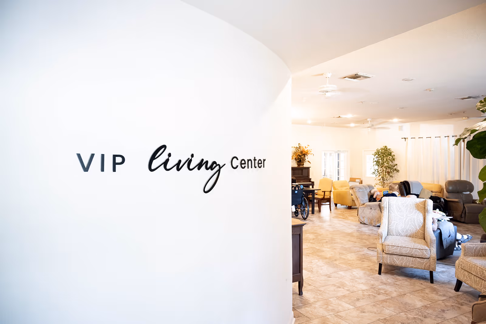 Curved white wall with a 'VIP living Center' sign opening into a bright communal lounge with armchairs and plants.