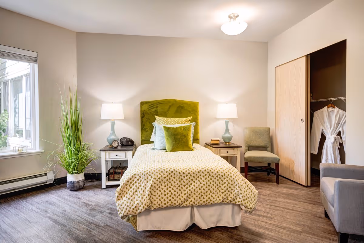 A cozy bedroom with a single bed featuring a green velvet headboard and patterned bedding. Two bedside tables with matching lamps flank the bed. A green chair is placed near a closet with sliding doors, one of which is open showing a white robe hanging inside. A large potted plant sits near a window letting in natural light. The room has wood flooring and neutral-colored walls.