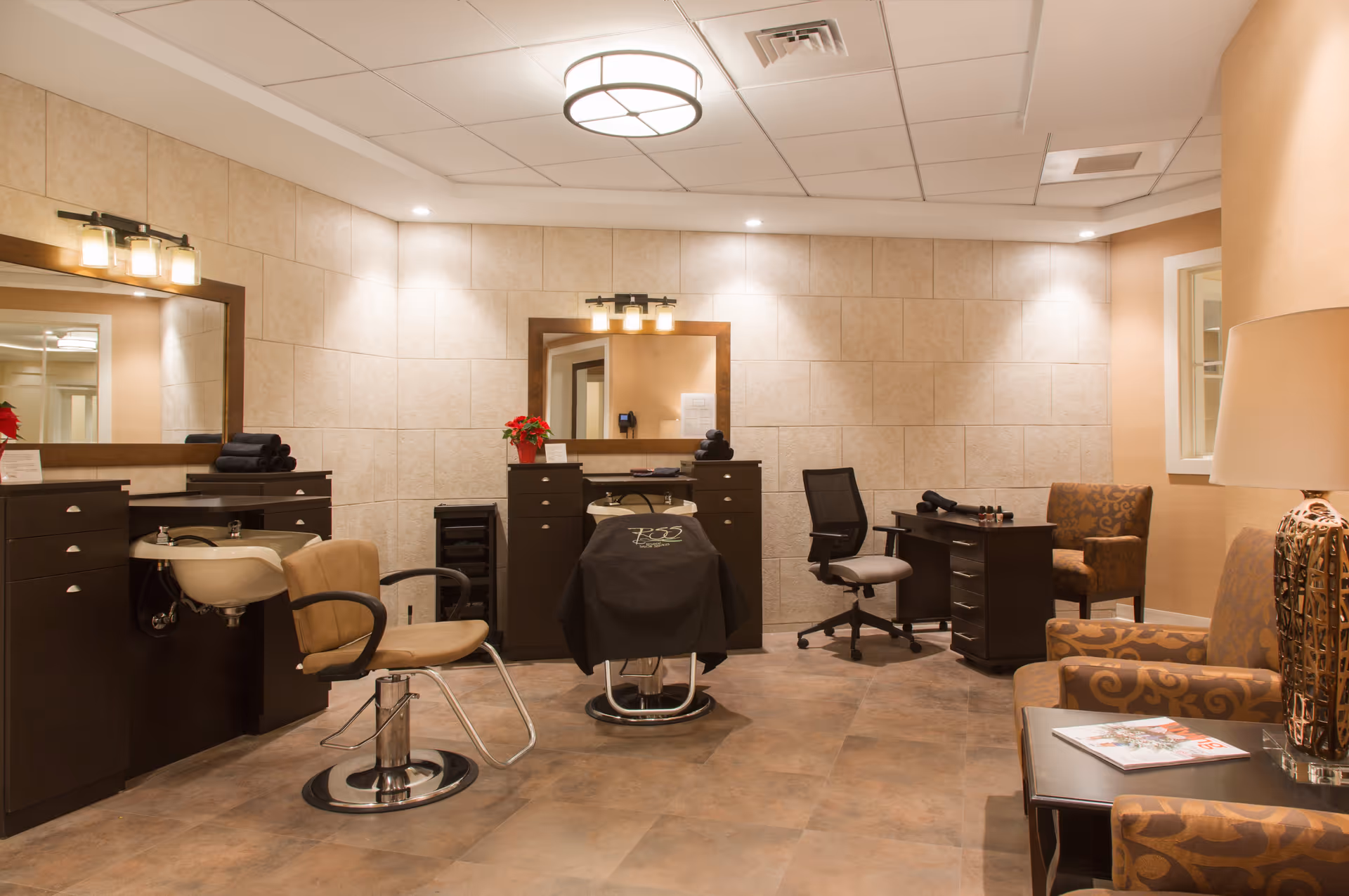 A well-lit salon room with two styling chairs in front of large mirrors, a sink for washing hair, a desk with a rolling chair, and comfortable armchairs with a side table and lamp. The walls are tiled and the floor has a warm-toned tile. There are small decorative plants and neatly folded towels on the counters.