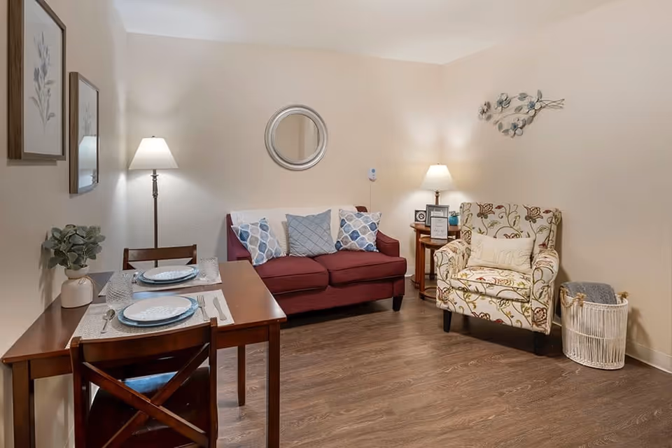 A cozy living area with a small wooden dining table set for two on the left, a maroon loveseat with three decorative pillows in the center, and a floral patterned armchair with a pillow on the right. The room has wooden flooring, two table lamps providing warm lighting, a round mirror on the wall above the loveseat, and framed floral artwork on the left wall. A basket with a blanket is placed next to the armchair.