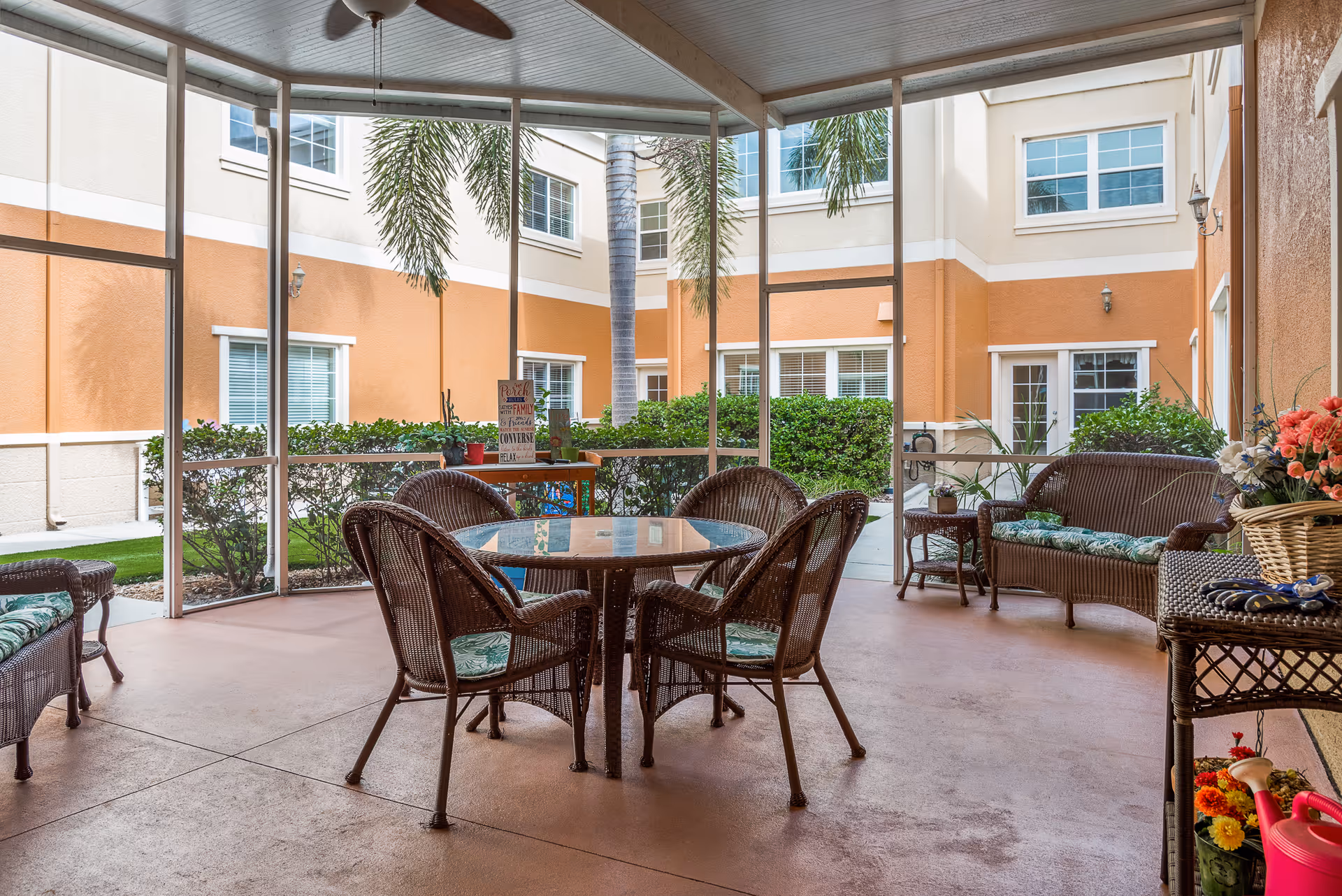 A screened-in patio area with a round glass-top table surrounded by four wicker chairs with green patterned cushions. There are two wicker loveseats with similar cushions, a small side table, and a wicker shelf holding a basket of flowers and gardening tools. The patio overlooks a courtyard with palm trees, bushes, and the exterior walls of a beige and orange building with multiple windows.