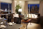 A cozy living room and dining area at night with large windows showcasing a city skyline view. The room features two brown sofas with cushions, a wooden coffee table, a dining table set with plates and glasses, a floor lamp, and a tall plant in the corner.