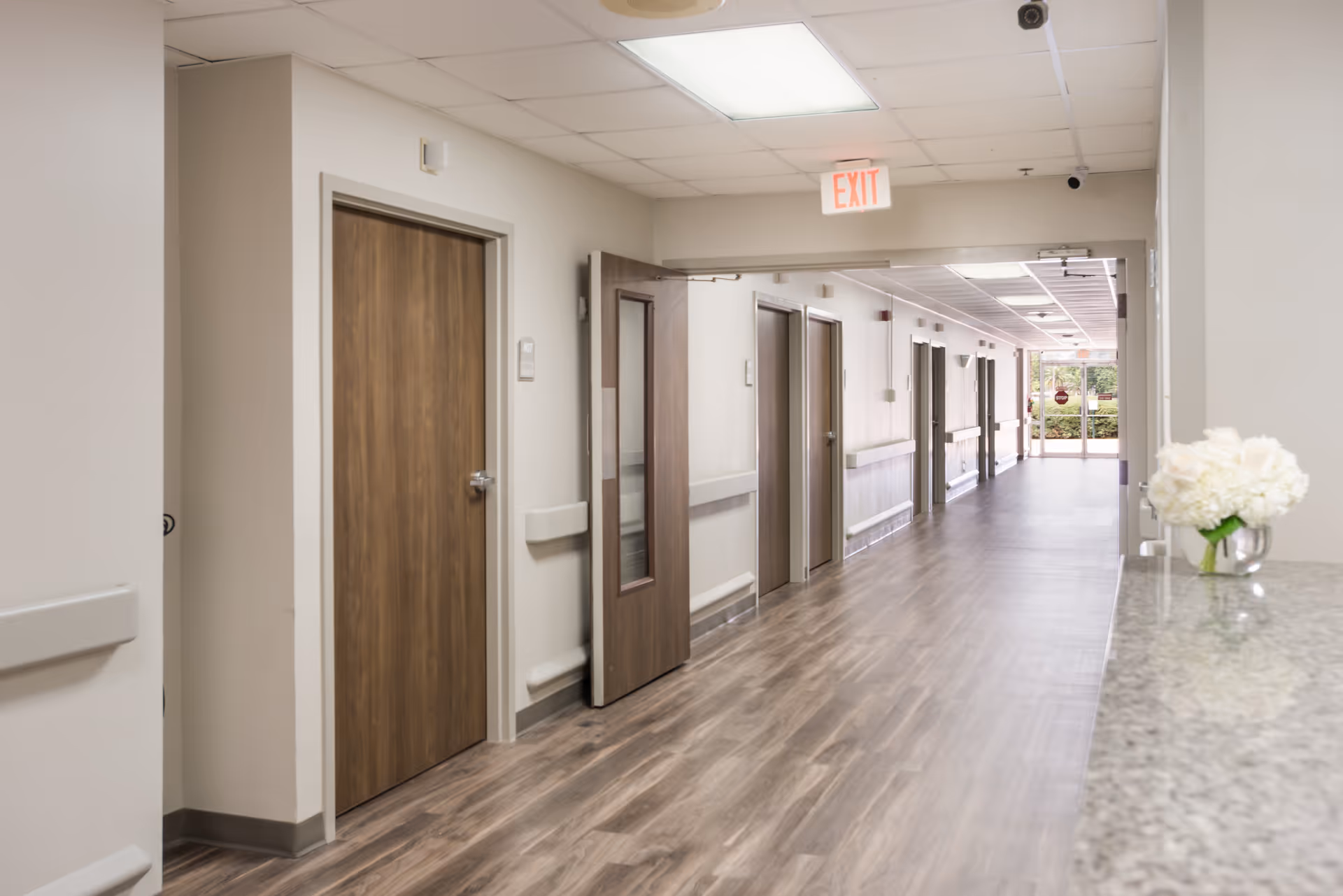 Bright interior hallway of a senior care facility with wood doors, handrails, an exit sign, and a reception counter with flowers.