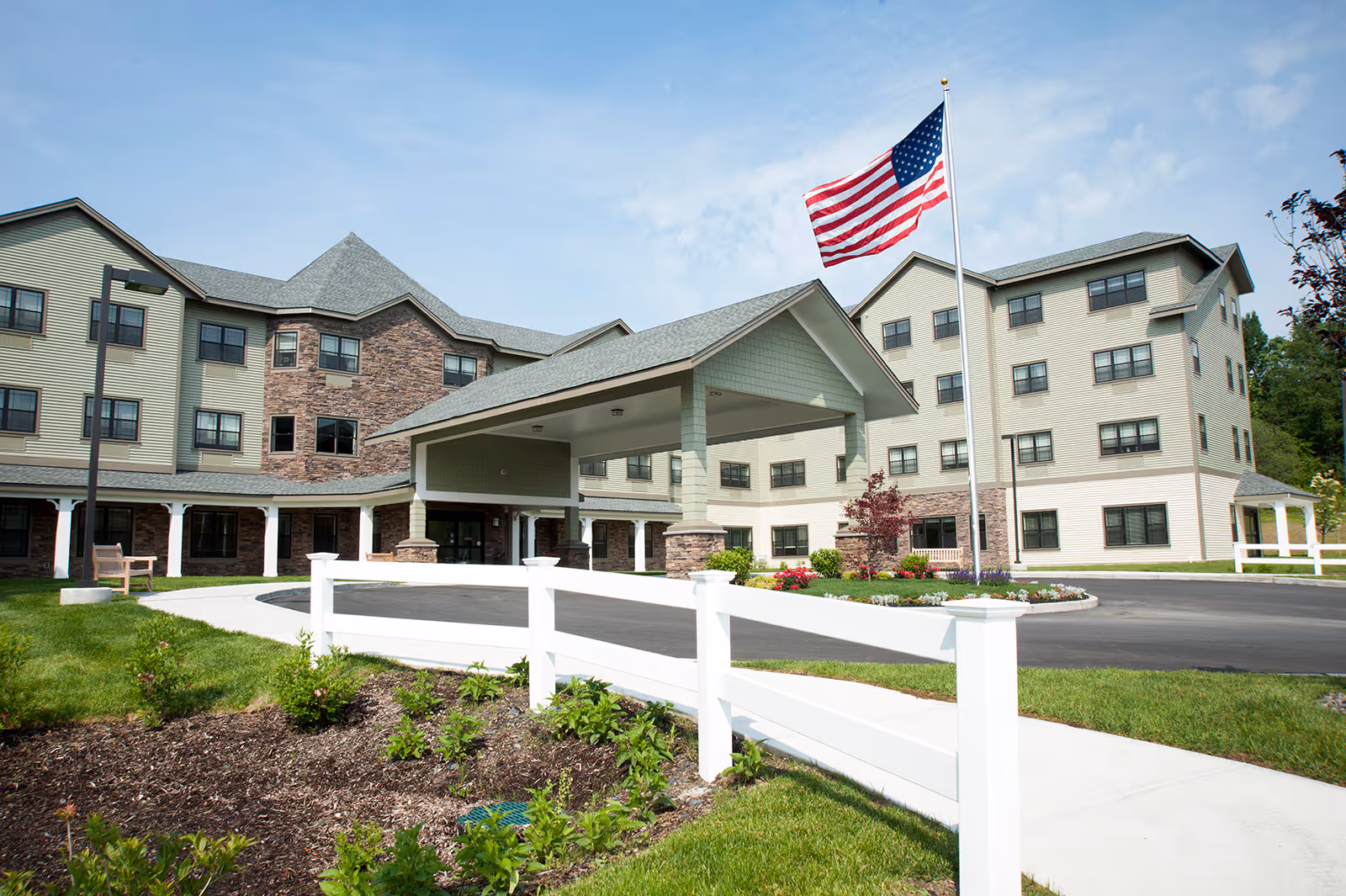Exterior view of Braemar at Wallkill senior living facility showing a multi-story building with a covered entrance, an American flag on a flagpole, landscaped garden beds, a white fence, and a clear blue sky.