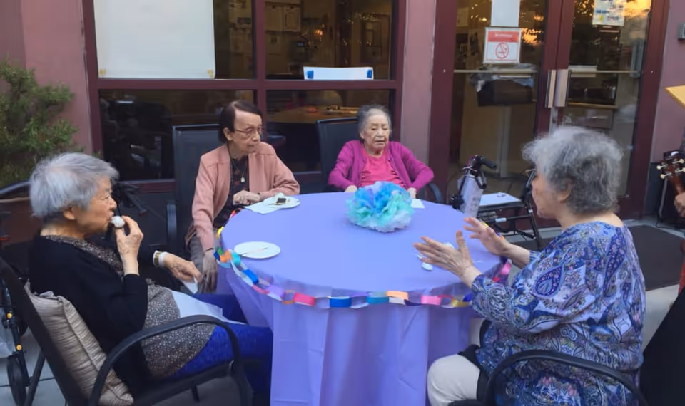 Four elderly women sitting around a round table covered with a purple tablecloth and decorated with a colorful paper chain and a blue and green tissue paper centerpiece. They are outdoors near a building entrance with glass doors and windows.
