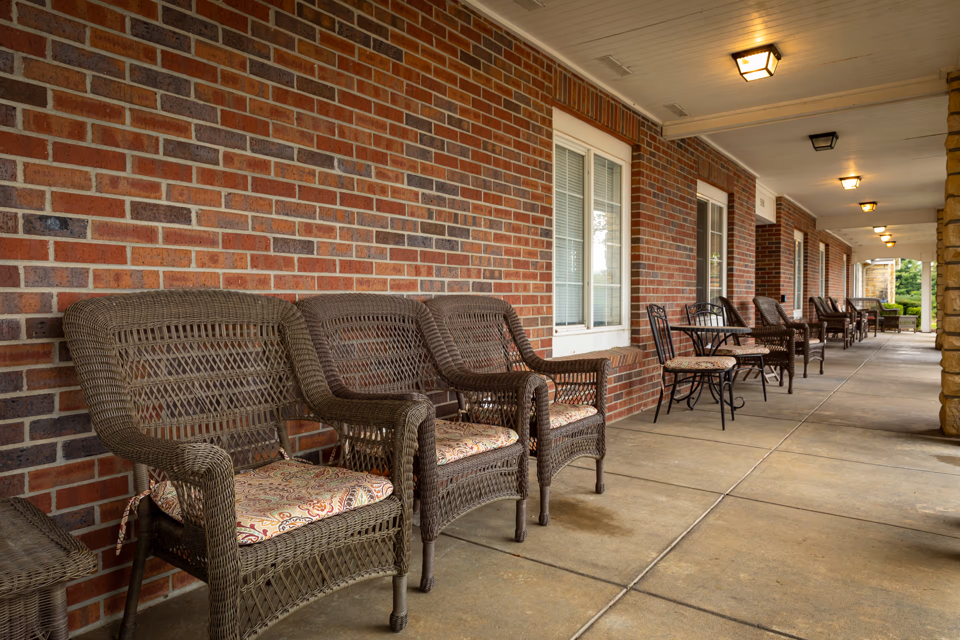 Covered brick porch with wicker chairs and small tables lined along a walkway.