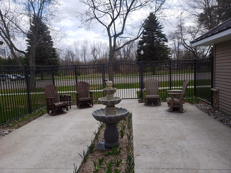 Outdoor patio area with a concrete floor, featuring a tiered stone fountain in the center surrounded by a narrow garden bed with small plants. There are four wooden Adirondack chairs placed around the patio, enclosed by a black metal fence with a gate. Trees and a road are visible in the background under a cloudy sky.