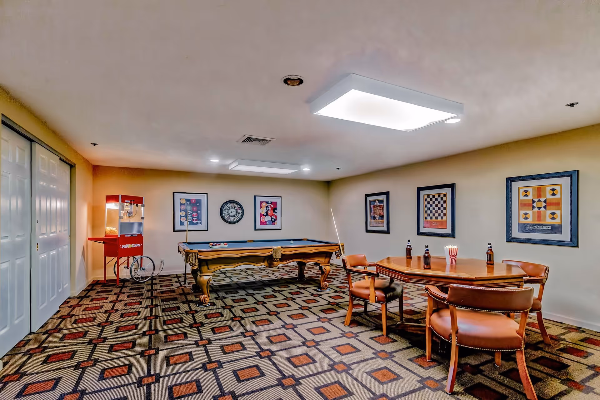 A recreational room with a pool table, a popcorn machine, and a wooden table surrounded by four brown leather chairs. The walls are decorated with framed artwork and a clock. The floor has a patterned carpet with geometric shapes.