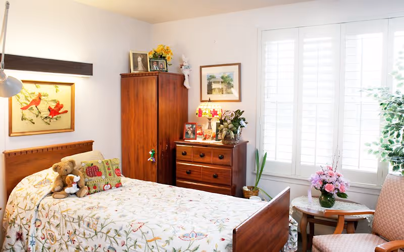 Sunlit bedroom with a wooden bed covered in a floral bedspread, stuffed animals, a wooden dresser and wardrobe, and a window with white shutters.