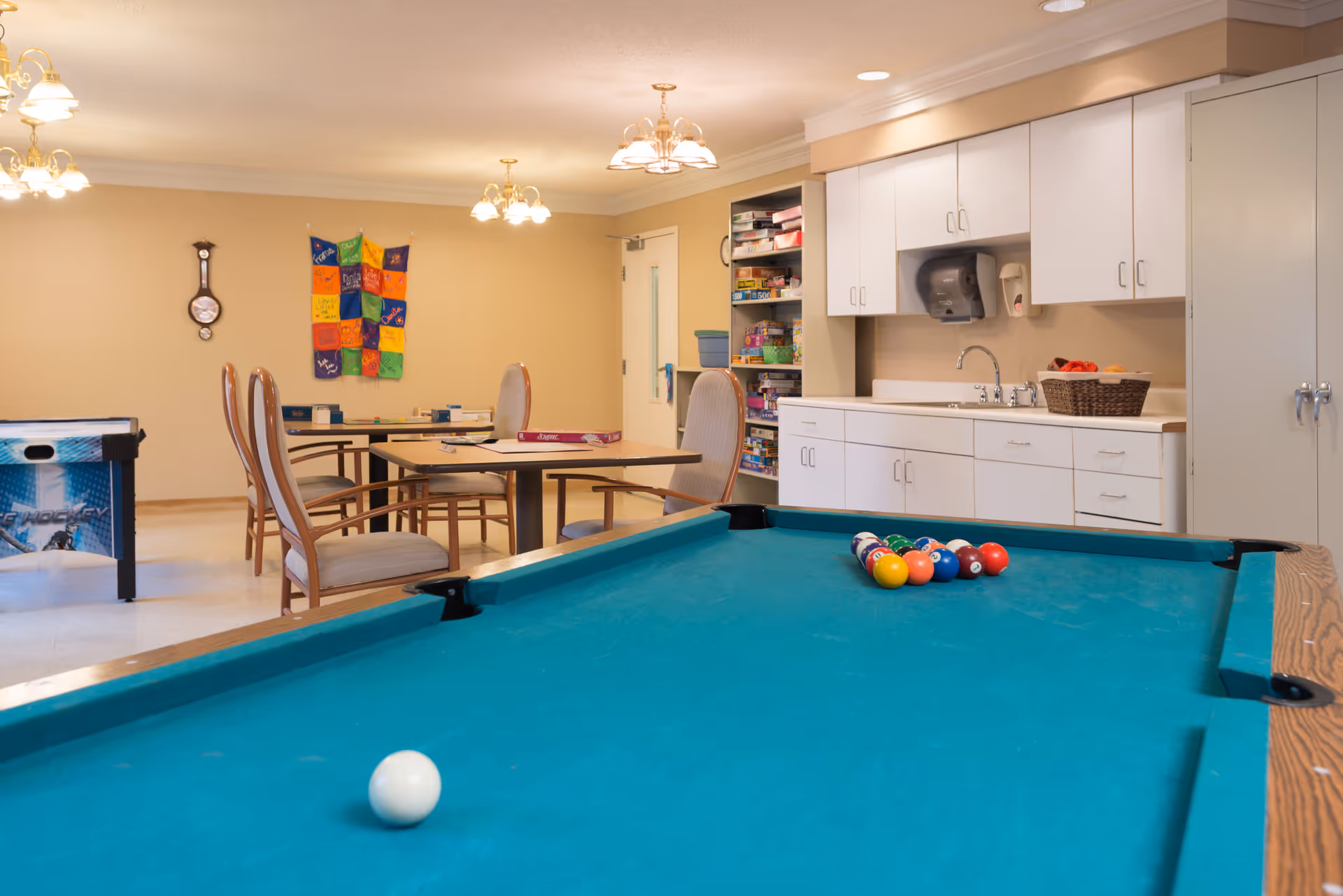 Recreation room featuring a pool table in the foreground, tables and chairs, a kitchenette with cabinets and sink, and shelves of games.