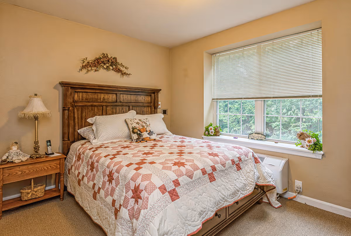 A cozy bedroom with a wooden headboard bed covered with a quilted bedspread featuring a star pattern in shades of red and white. There are several pillows on the bed, including a decorative one with a floral design. To the left of the bed is a wooden nightstand with a vintage-style lamp, a small doll, and a phone. A large window with white blinds partially open lets in natural light and shows greenery outside. The window sill has small decorative plants and a 'Welcome' sign.