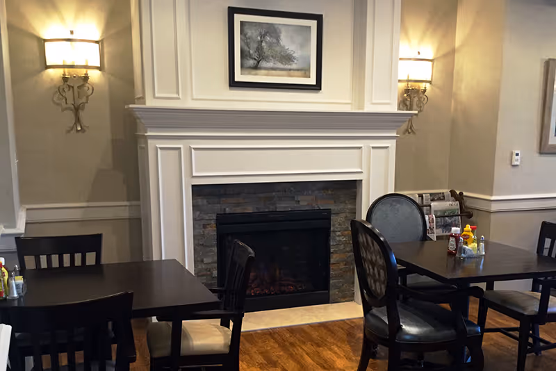Interior view of a cozy dining area with dark wooden tables and chairs, a white fireplace with a stone surround, a framed picture above the fireplace, and wall sconces providing warm lighting.