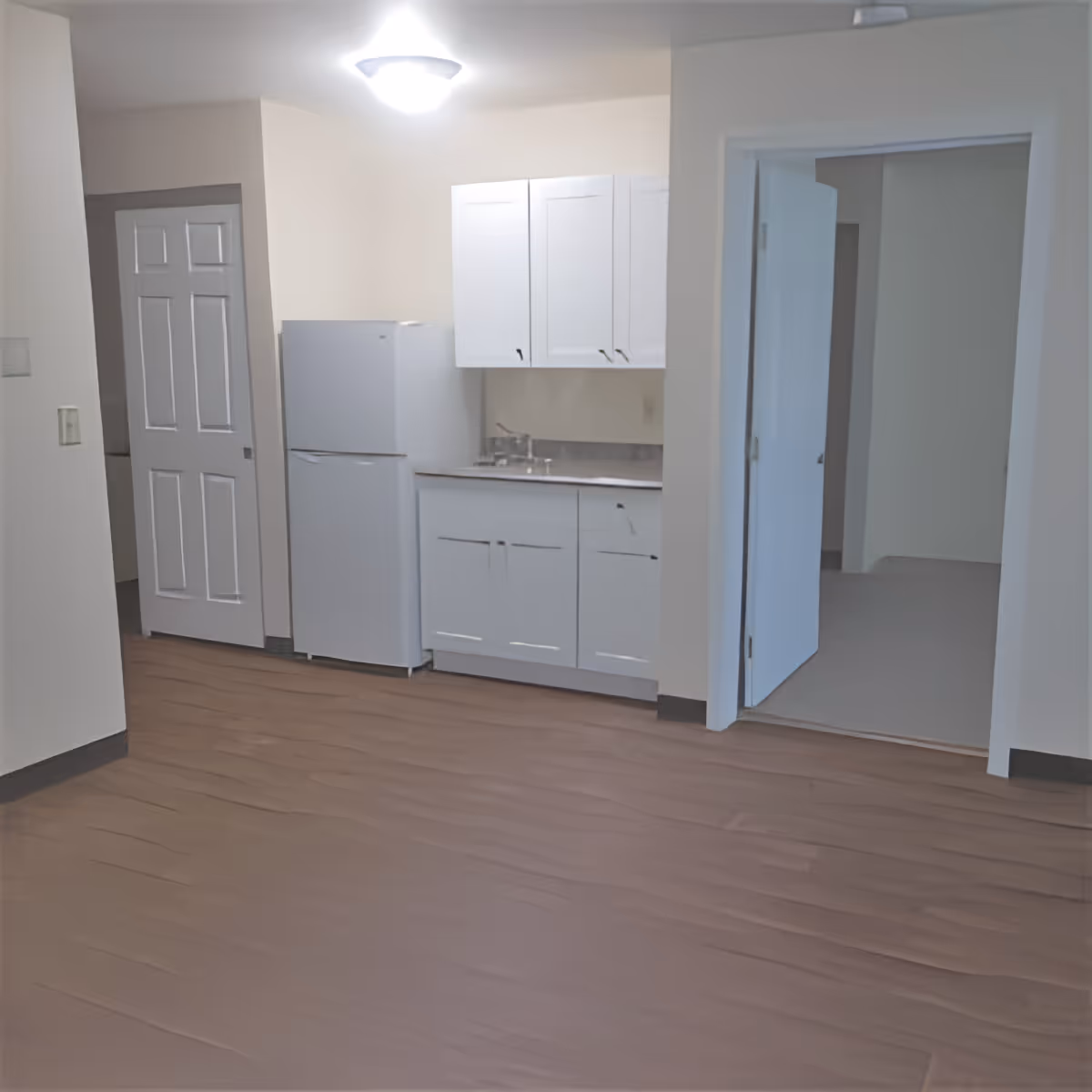 A small kitchen area with white cabinets, a white refrigerator, and a sink. The floor is wooden, and there are two doorways visible, one with a closed white door and the other open leading to another room.