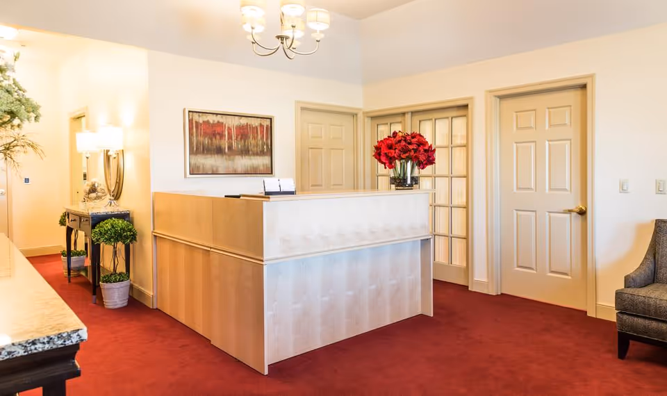 Reception area with a light wood front desk adorned with a vase of red flowers. The room has beige walls, red carpet flooring, a chandelier, and several doors including one with glass panels. There is a framed abstract painting on the wall and a small table with a lamp and plants near the entrance.