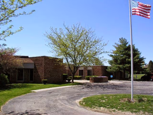 Exterior view of a single-story brick building with a curved driveway, green grass, trees, and an American flag on a flagpole in front of the building under a clear blue sky.