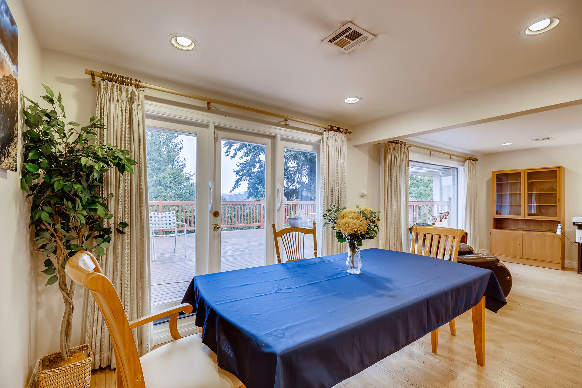 A dining area with a wooden table covered by a blue tablecloth and a vase of yellow flowers in the center. There are wooden chairs around the table. Large glass sliding doors with cream-colored curtains open to a wooden deck outside. The room has light-colored wooden flooring, recessed ceiling lights, a potted plant in the corner, and a wooden cabinet with glass doors in the background.