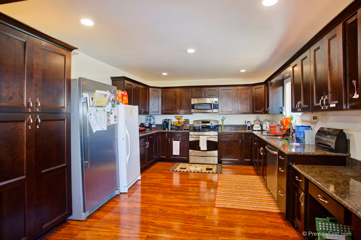 A spacious kitchen with dark wooden cabinets, stainless steel appliances including a refrigerator, oven, microwave, and dishwasher. The kitchen has granite countertops, wooden flooring, and recessed ceiling lights. Various kitchen items such as a toaster, blender, coffee maker, and fruit bowl are visible on the counters.