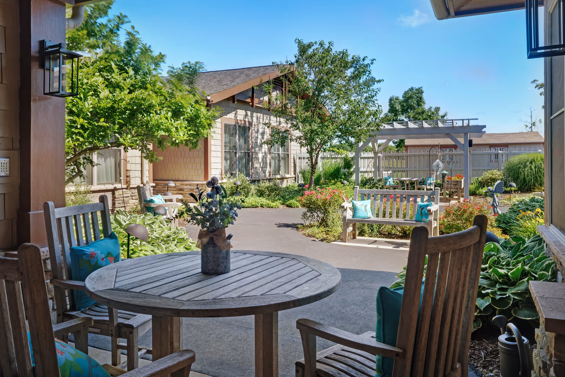 Covered patio with a round wooden table and chairs opening onto a landscaped courtyard with benches, a pergola, and surrounding building.