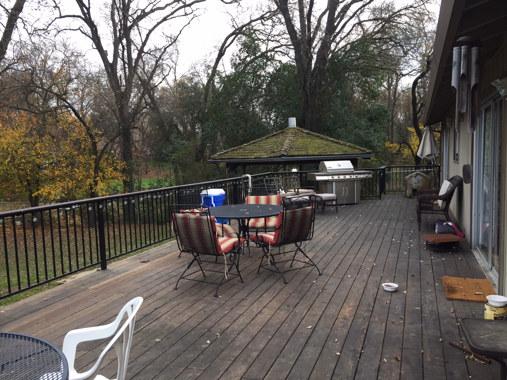 Outdoor wooden deck with several chairs and tables, a barbecue grill, a cooler, and a small gazebo with a moss-covered roof in the background. The deck is surrounded by a black metal railing and overlooks a wooded area with trees showing autumn foliage.