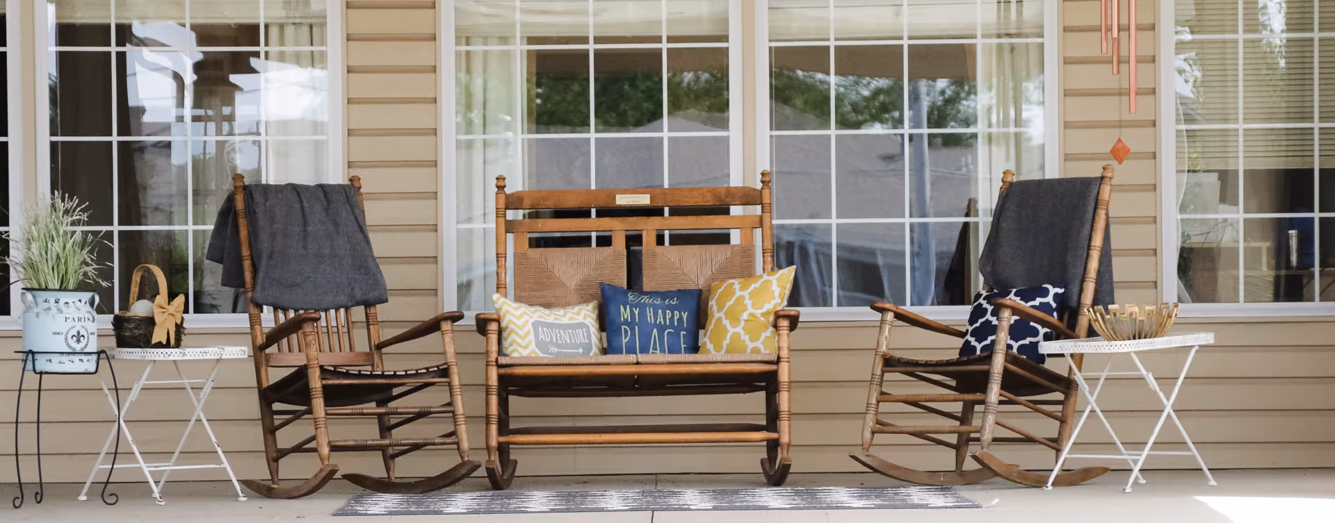 A covered front porch with three wooden rocking chairs (including a double bench) decorated with pillows, small side tables and a potted plant.