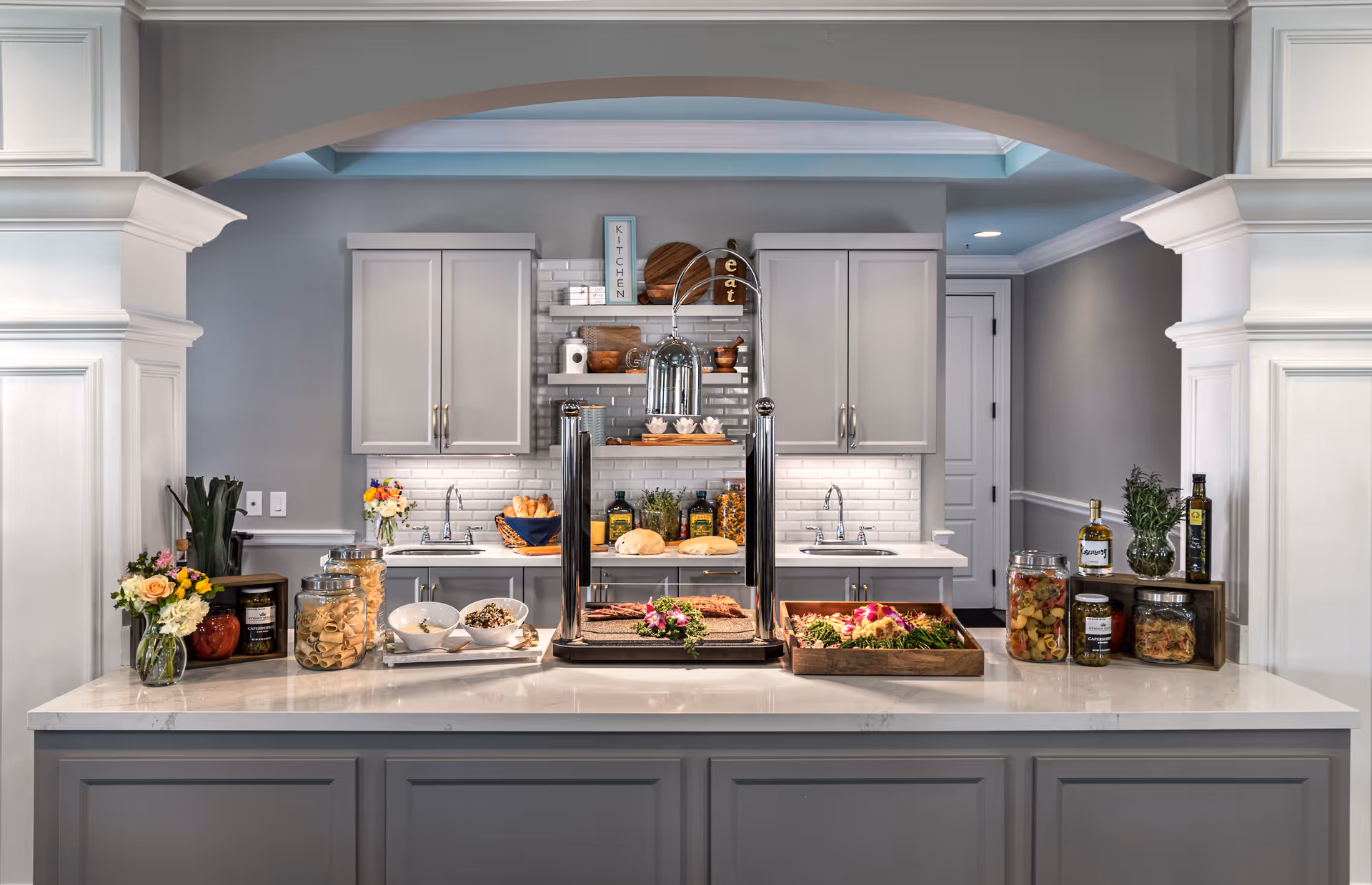 A light-gray kitchen serving counter with cabinets, sinks, food display trays, jars, and floral arrangements under an arched pass-through.