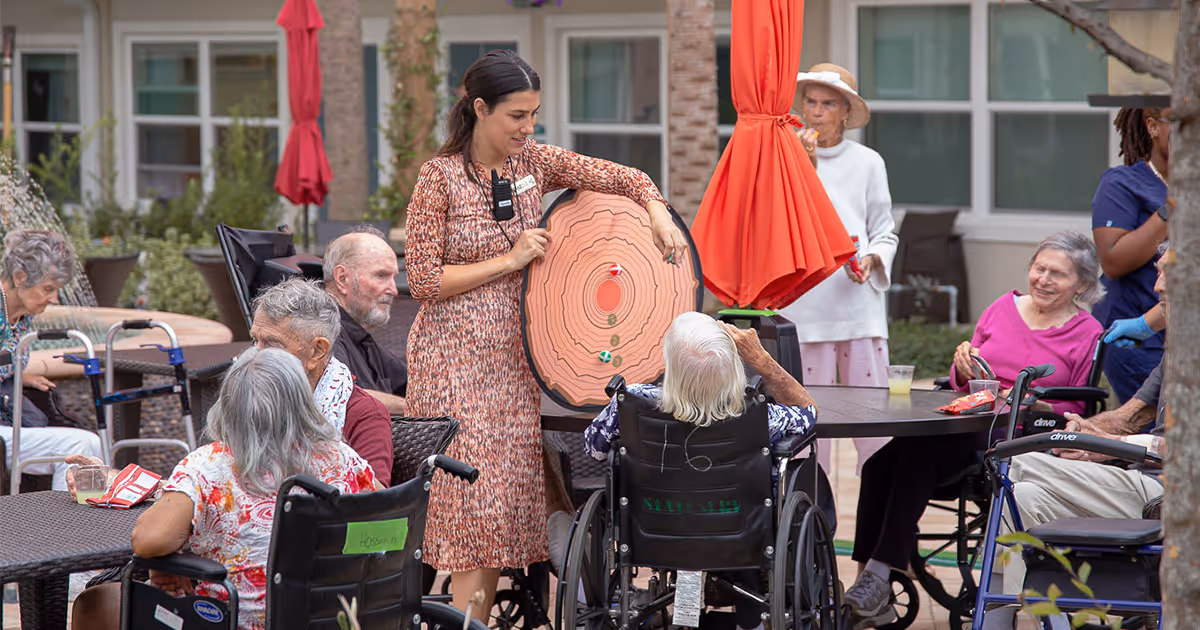 A group of elderly residents, some in wheelchairs, gathered outdoors around tables with a staff member holding a large target game board. The setting appears to be a courtyard or patio area of an assisted living facility, with umbrellas and building windows in the background.