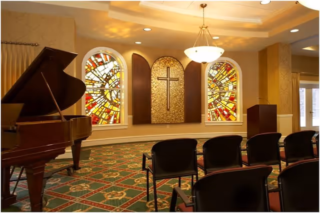 Interior room with a grand piano on the left, rows of chairs facing a wall with two stained glass windows and a central panel featuring a cross. The room has patterned carpet, warm lighting, and a chandelier hanging from the ceiling.