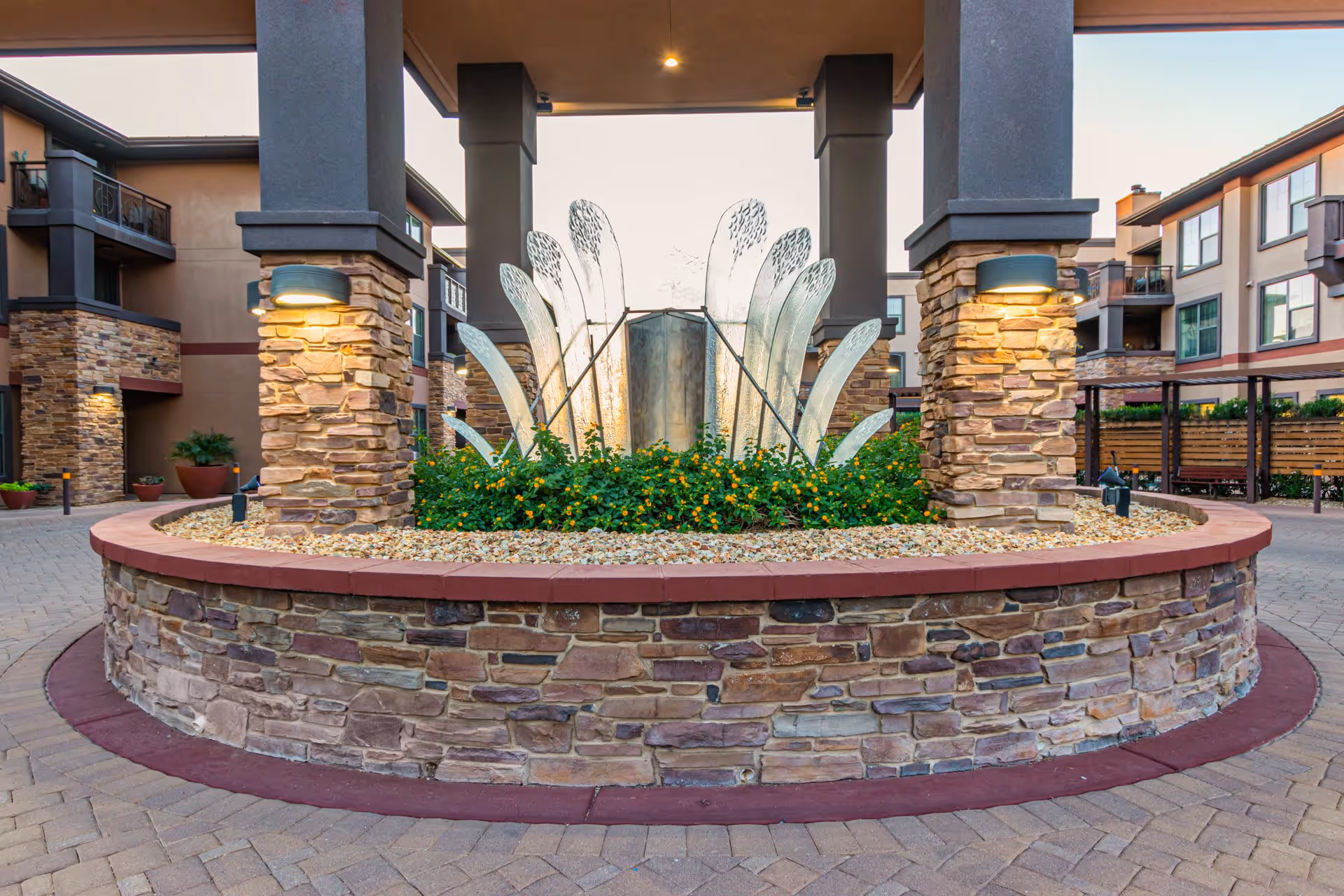Outdoor courtyard area with a circular stone planter filled with green plants and yellow flowers, featuring a modern metal sculpture resembling abstract leaves or petals. The planter is surrounded by stone pillars with mounted lights, and residential buildings with balconies are visible in the background.