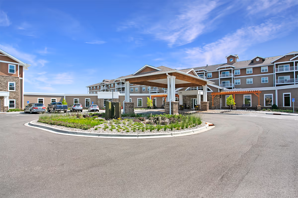 Front exterior view of Norris Square I facility showing a circular driveway with a landscaped island in the center, a covered entrance supported by stone pillars, multiple parked cars, and a multi-story building under a blue sky with some clouds.