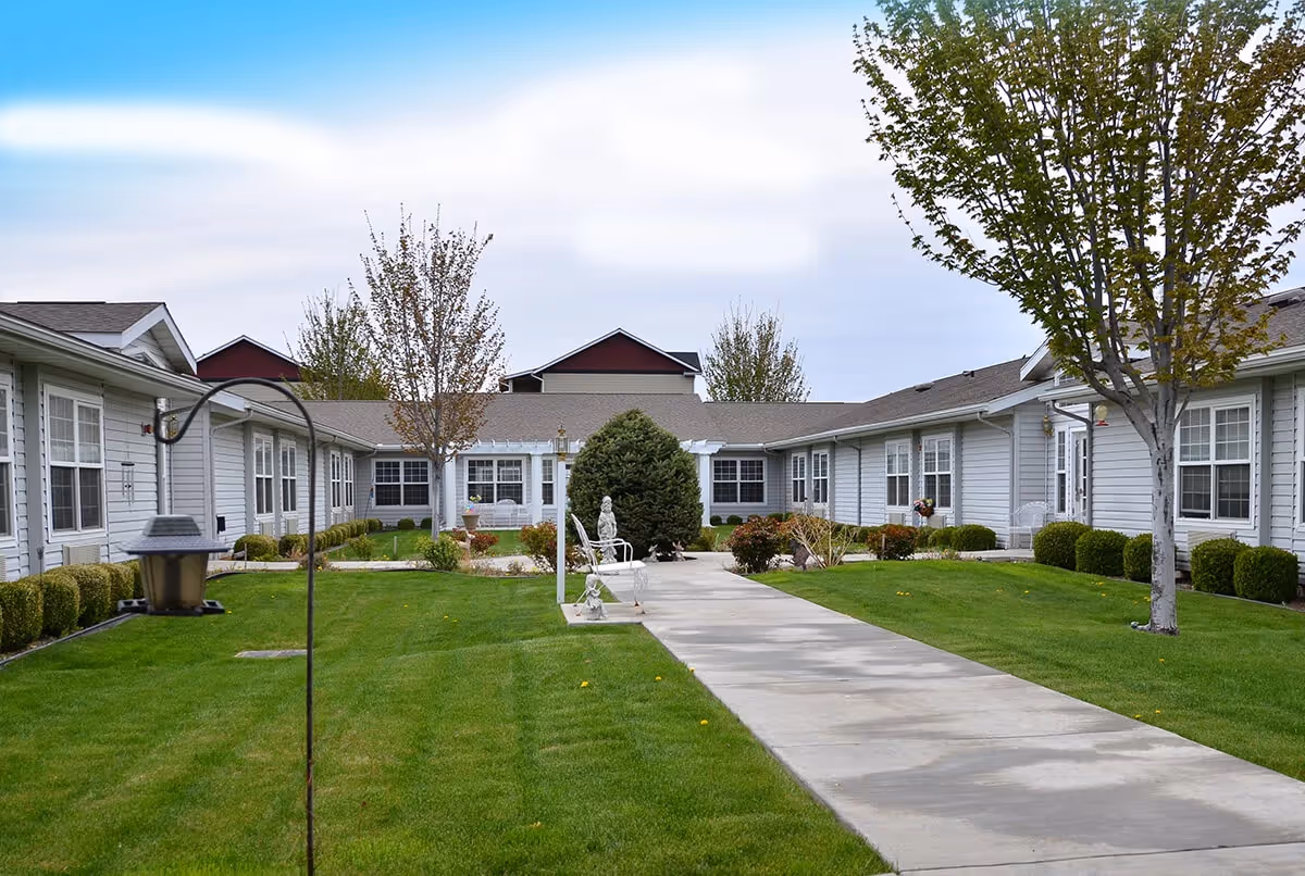 Outdoor courtyard area of Avista Senior Living Yakima with a concrete walkway, green grass, trees, bushes, and benches surrounded by single-story residential buildings with multiple windows.