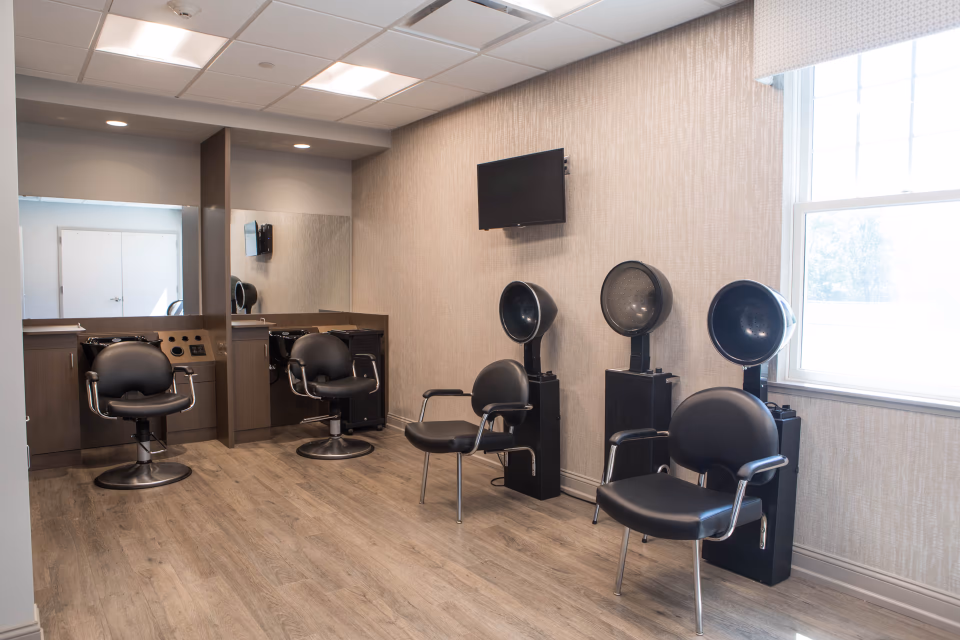 Interior view of a hair salon area in a senior living facility with black salon chairs, hair drying stations, a wall-mounted TV, large mirror, and a window letting in natural light.