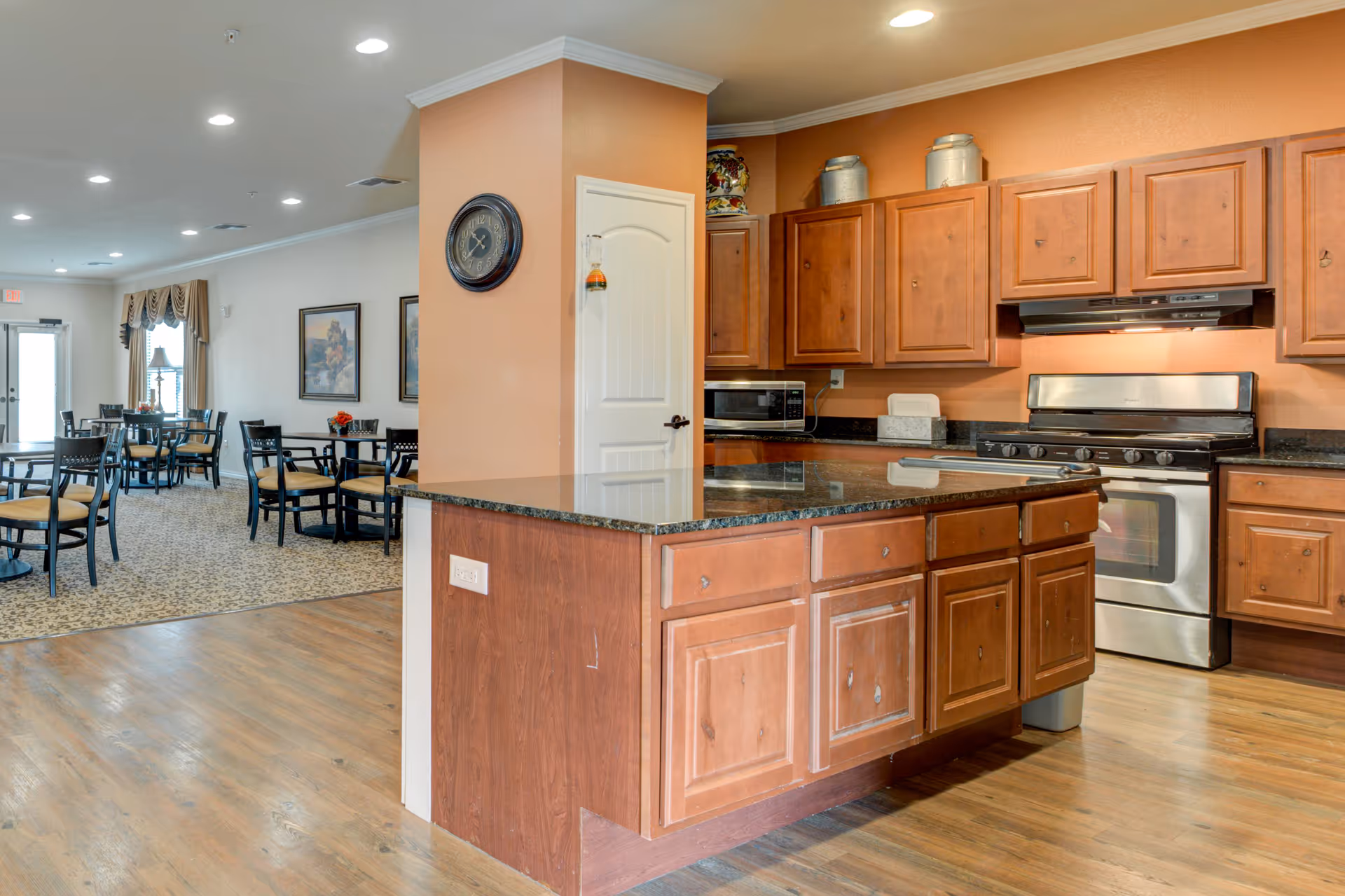 Interior view of a senior living facility showing a kitchen area with wooden cabinets, a stove, microwave, and a large island with a granite countertop. Adjacent to the kitchen is a dining area with multiple tables and chairs, carpeted flooring, and framed artwork on the walls.