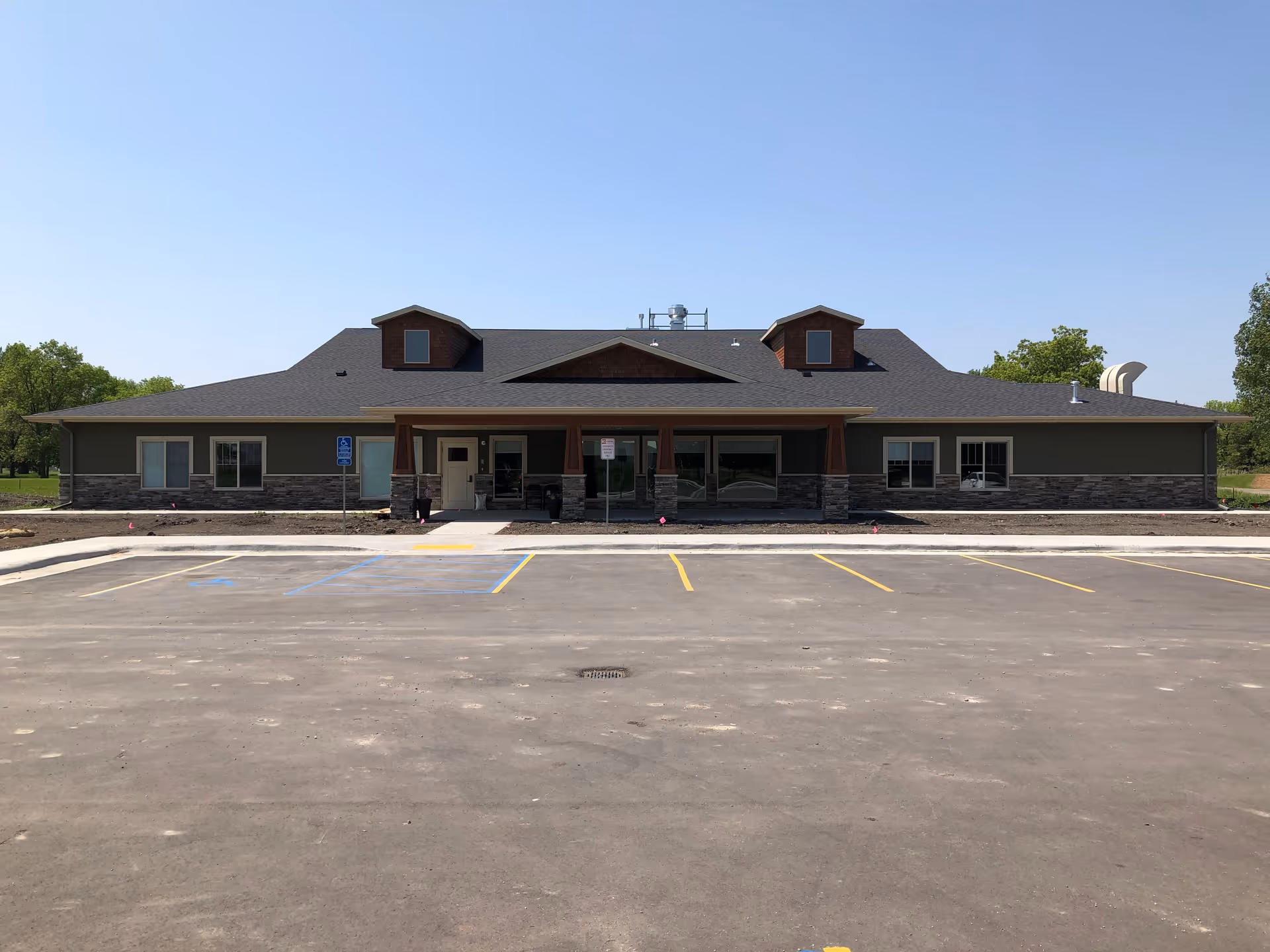 Front exterior view of a single-story assisted living facility building with a dark roof, stone accents on the lower walls, and a covered entrance. The building is surrounded by a paved parking lot with marked parking spaces and a clear blue sky overhead.