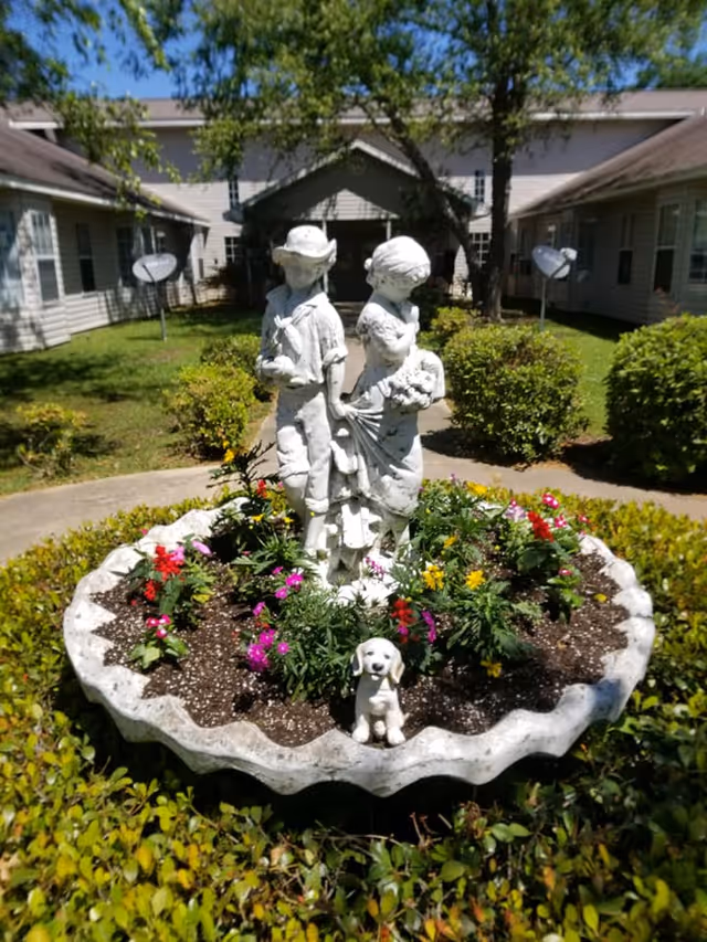 A garden area with a circular flower bed containing colorful flowers and a white statue of a boy and girl standing back to back, along with a small dog statue at the front. The garden is surrounded by bushes and pathways, with a building and trees in the background under a clear blue sky.